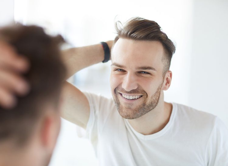 Portrait of handsome young man with lush hair and short stubble looking at his reflection in mirror and smiling, copy space
