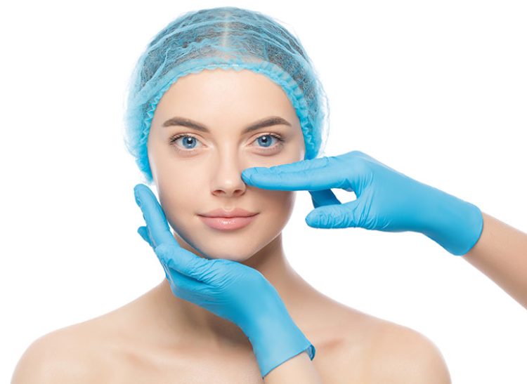 Young woman ready for rhinoplasty, doctor in blue gloves touching her nose, isolated on white background